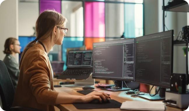 woman viewing code on two monitors with coworker seated to her left